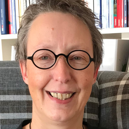 Eine Person mit kurzen Haaren, einer runden Brille und einem breiten Lächeln vor einem Bücherregal.