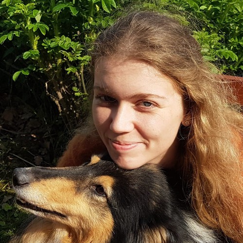 Eine Person mit langen, dunkelblonden, welligen Haaren schaut verschmitzt in die Kamera. Der Hintergrund ist aus grünen Blättern. Am unteren Ende ist ein Hundekopf zu sehen, die Person stützt ihr Kinn auf den Kopf des Hundes, der zu lächeln scheint.